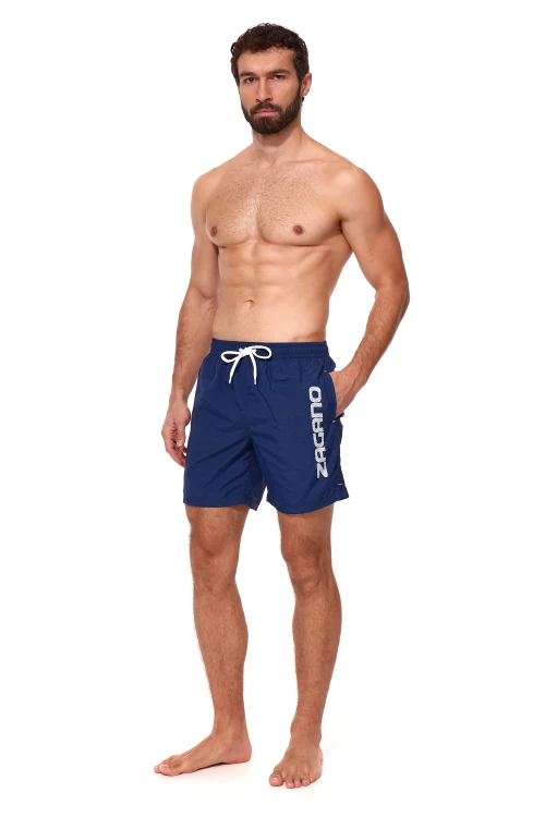 Granatowe długie szorty kąpielowe męskie ZAGANO 5115 LONG-NAVY BLUE z białym sznurkiem do regulacji i dużym pionowym logo marki na nogawce. Wygodny, sportowy fason z bocznymi kieszeniami i tylną kieszenią z klapką, idealny na basen, plażę i do pływania.